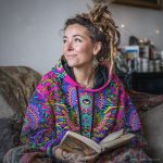 Alternative looking hippy style woman sitting down inside and boho home, wearing an Alex Tooth Radiant Bloom Sanctum Hoodie.