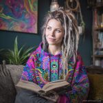 Alternative looking hippy style woman sitting down inside and boho home, wearing an Alex Tooth Radiant Bloom Sanctum Hoodie.