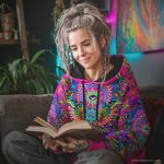 Alternative looking hippy style woman sitting down inside and boho home, wearing an Alex Tooth Radiant Bloom Sanctum Hoodie.