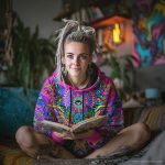 Alternative looking hippy style woman sitting down inside and boho home, wearing an Alex Tooth Radiant Bloom Sanctum Hoodie.
