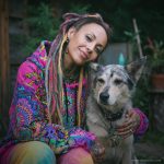 Woman wearing Radiant Bloom Sanctum Hoodie by Alex Tooth with dreadlocks, tattoos, and colourful clothing posing with a dog outdoors.