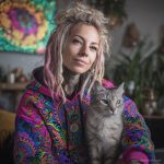 Alternative looking hippy style woman sitting down inside and boho home, wearing an Alex Tooth Radiant Bloom Sanctum Hoodie.