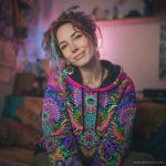 Alternative looking hippy style woman sitting down inside and boho home, wearing an Alex Tooth Radiant Bloom Sanctum Hoodie.