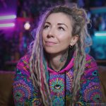 Alternative looking hippy style woman sitting down inside and boho home, wearing an Alex Tooth Radiant Bloom Sanctum Hoodie.