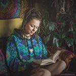 Alternative looking hippy style woman sitting down inside and boho home, wearing an Alex Tooth Emerald StarGate Sanctum Hoodie.