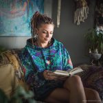 Alternative looking hippy style woman sitting down inside and boho home, wearing an Alex Tooth Emerald StarGate Sanctum Hoodie.