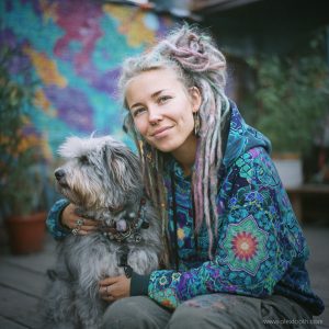 Colourful dreadlocked woman with a grey dog in front of a painted mural — visionary art by Alex Tooth.