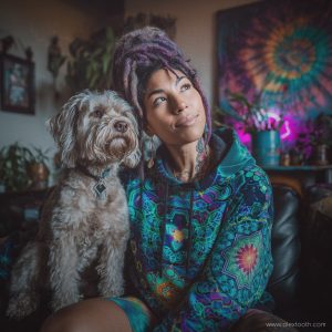 Vibrant woman with dreadlocks and tattoos sitting with dog indoors, colourful psychedelic art in background.