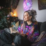 Alternative looking hippy style woman sitting down inside and boho home, wearing an Alex Tooth Inner Flight Sanctum Hoodie.