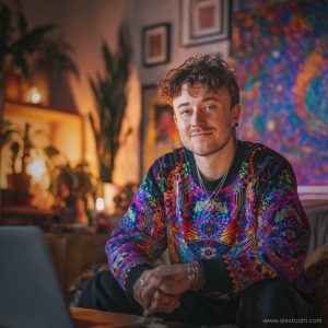 Brightly lit portrait of a young man indoors, wearing a colourful, psychedelic-patterned sweatshirt, with artwork and plants in the background.