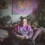 Cool looking alternative hippy style man sitting in a boho home reading a book wearing Alex Tooth Radiant Bloom Stillpoint Sweatshirt.