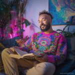 Cool looking alternative hippy style man sitting in a boho home reading a book wearing Alex Tooth Radiant Bloom Stillpoint Sweatshirt.