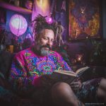 Cool looking alternative hippy style man sitting in a boho home reading a book wearing Alex Tooth Radiant Bloom Stillpoint Sweatshirt.