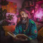 Cool looking alternative hippy style man sitting in a boho home reading a book wearing Alex Tooth Emerald StarGate Stillpoint Sweatshirt.