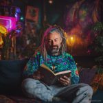 Cool looking alternative hippy style man sitting in a boho home reading a book wearing Alex Tooth Emerald StarGate Stillpoint Sweatshirt.