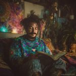 Cool looking alternative hippy style man sitting in a boho home reading a book wearing Alex Tooth Emerald StarGate Stillpoint Sweatshirt.
