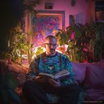 Cool looking alternative hippy style man sitting in a boho home reading a book wearing Alex Tooth Emerald StarGate Stillpoint Sweatshirt.