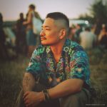 Cool looking hippy type alternative style man sitting around a campfire wearing and Alex Tooth Emerald StarGate Island Flow Hawaiian Shirt.