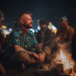 Cool looking hippy type alternative style man sitting around a campfire wearing and Alex Tooth Emerald StarGate Island Flow Hawaiian Shirt.