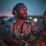 Cool looking hippy type alternative style man sitting around a campfire wearing and Alex Tooth Emerald StarGate Island Flow Hawaiian Shirt.