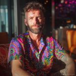 Cool looking hippy type alternative style man sitting inside wearing and Alex Tooth Radiant Bloom Island Flow Hawaiian Shirt.