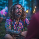 Cool looking hippy type alternative style man sitting around a campfire wearing and Alex Tooth Radiant Bloom Island Flow Hawaiian Shirt.