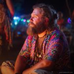 Cool looking hippy type alternative style man sitting around a campfire wearing and Alex Tooth Radiant Bloom Island Flow Hawaiian Shirt.