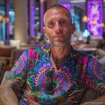 Cool looking hippy type alternative style man sitting inside wearing and Alex Tooth Radiant Bloom Island Flow Hawaiian Shirt.