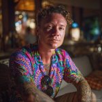 Cool looking hippy type alternative style man sitting inside wearing and Alex Tooth Radiant Bloom Island Flow Hawaiian Shirt.