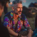 Cool looking hippy type alternative style man sitting around a campfire wearing and Alex Tooth Inner Flight Island Flow Hawaiian Shirt.