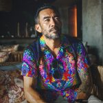 Cool looking hippy type alternative style man sitting inside wearing and Alex Tooth Inner Flight Island Flow Hawaiian Shirt.