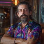 Cool looking hippy type alternative style man sitting inside wearing and Alex Tooth Inner Flight Island Flow Hawaiian Shirt.
