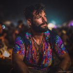 Cool looking hippy type alternative style man sitting around a campfire wearing and Alex Tooth Inner Flight Island Flow Hawaiian Shirt.