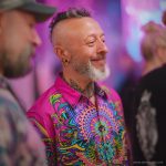 Smart looking alternative hippy style man standing in a inside social function setting wearing an Alex Tooth Radiant Bloom InnerLight Shirt.