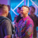 Smart looking alternative hippy style man standing in a inside social function setting wearing an Alex Tooth Radiant Bloom InnerLight Shirt.
