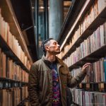 A man reading a book in a library aisle, surrounded by shelves of books wearing Alex Tooth Inner Flight Long Sleeved Balance Tee T-Shirt.