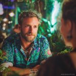 Smart looking alternative hippy style man sitting inside a boho cafe / restaurant setting wearing an Alex Tooth Emerald StarGate InnerLight Shirt.