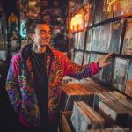Alternative hippy looking man standing inside a record store wearing Alex Tooth Radiant Bloom Pathfinder Jacket.