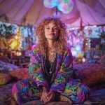 Alternative looking hippy woman sitting outside smiling, wearing long zip up fleece lined Alex Tooth Radiant Bloom Ritual Robe