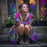 Alternative looking hippy woman sitting outside smiling, wearing long zip up fleece lined Alex Tooth Radiant Bloom Ritual Robe